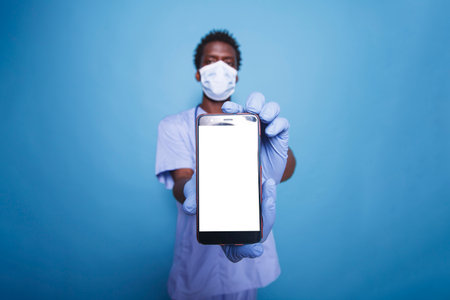 African American doctor stands confidently in scrubs, holding a mobile device with a blank white screen. Male medical assistant vertically holding smartphone with isolated copyspace mockup template.の写真素材