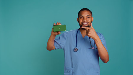 Portrait of joyous male nurse pointing finger towards isolated screen smartphone. Radiant healthcare specialist holding chroma key phone, blue studio background, camera Bの写真素材