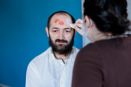 Girlfriend helping man prepare for halloween party, dressing him as terrifying creepy zombie. Woman applying scary makeup on boyfriend, turning him into undead monster with scarsの写真素材