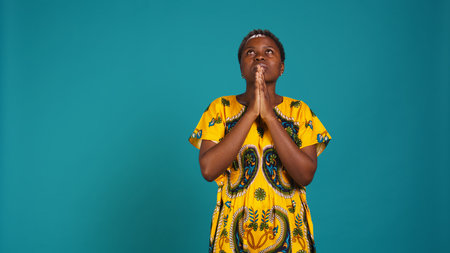 Spiritual hopeful woman holding hands in a prayer and having belief in Jesus, asking for luck and good health against blue background. Young adult believing in spirituality and religion. Camera B.の写真素材