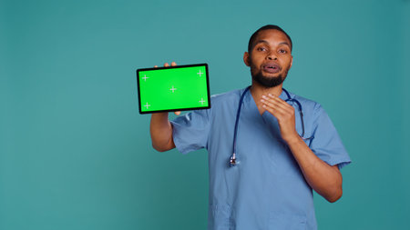 Portrait of african american male nurse showing medical instructions video on isolated screen tablet. Healthcare worker holding mockup digital device, studio backdrop, camera Bの写真素材