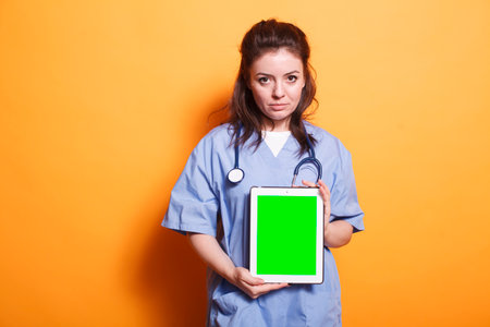 Healthcare professional vertically holding tablet with green screen. Woman wearing nurse uniform and stethoscope is grasping digital device displaying blank chromakey mockup template.の写真素材
