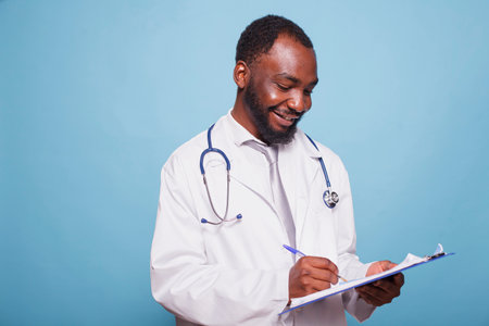 Smiling black doctor dressed in white lab coat and stethoscope standing with a clipboard containing medical records. Cheerful african american physician in hospital uniform writes on patient charts.の写真素材