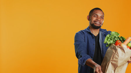 Vegan adult presenting his ripe freshly harvested produce in a paper bag, recommending local farming and sustainable lifestyle. Male model feeling pleased with his organic groceries. Camera B.の写真素材