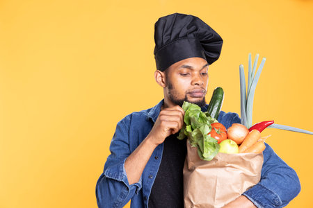 Young professional chef smelling fresh fruits and vegetables in a paper bag, preparing to cook a vegan healthy meal recipe. African american guy enjoys eating natural produce.の写真素材