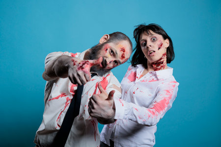 Couple doing professional photoshoot dressed as zombies for Halloween party, isolated over blue studio background. Man and woman covered in fake blood and scars pretending to be ghoulsの写真素材
