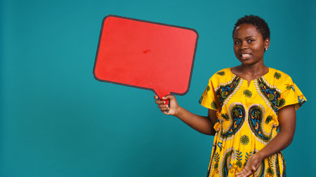 Smiling person showing a red isolated speech bubble in studio, holding a cardboard icon for a commercial advertisement. Young woman in tribal attire presenting an empty carton sign. Camera B.の写真素材