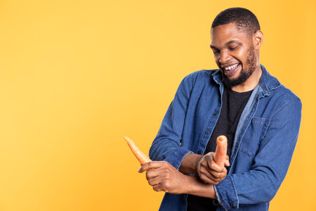 Charismatic african american guy pretending to play drums with carrots, having fun with veggies against yellow background. Funny person fooling around with bio produce in studio.の写真素材