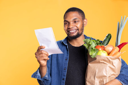 Young adult smiling while he verifies his shopping list on camera, buying all needed ingredients for a vegan recipe. Cheerful confident african american guy looks for ripe fruits and veggies.の写真素材