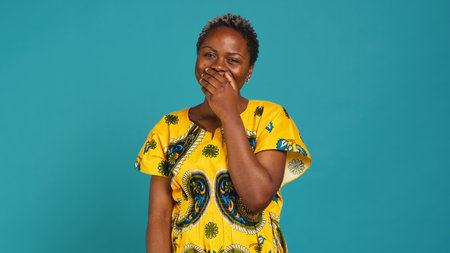 Cute female model giggling against blue background in studio, wearing floral pattern dress to symbolize tribal ethnic culture. Happy confident girl laughing, natural beauty. Camera A.の写真素材
