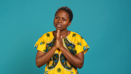 Confident optimistic woman holding hands in a prayer and talks to Jesus, praying for good luck and health against blue background. Young adult with spiritual belief being religious. Camera A.の写真素材