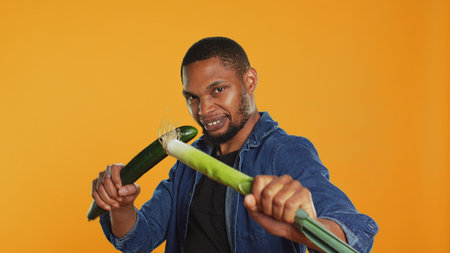 Pleased young adult pretending to start a fight with a cucumber and a leek, acting silly having fun in studio. Vegan guy playing around with natural eco friendly green vegetables. Camera A.の写真素材