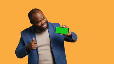 Portrait of man doing influencer marketing using isolated screen phone, showing thumbs up sign, studio background. Merry BIPOC person holding empty copy space mockup cellphone, camera Aの写真素材