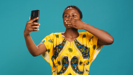 Charming natural woman posing with confidence for a selfie, taking pictures with a smartphone against blue background. Young smiling person takes photographs in studio. Camera A.の写真素材
