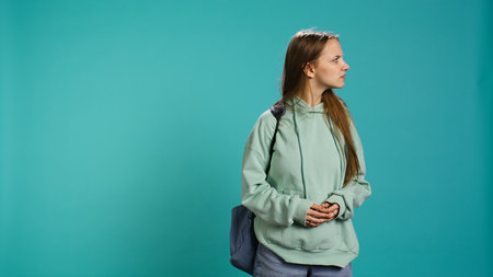 Young woman holding backpack looking around for something, doing hand gesture, isolated over studio background. Radiant teenager with good eyesight seeking things, camera Bの写真素材