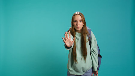 Assertive young woman doing stop hand gesture and talking. Annoyed girl doing firm halt sign gesturing, wishing to end concept, isolated over blue studio background, camera Bの写真素材