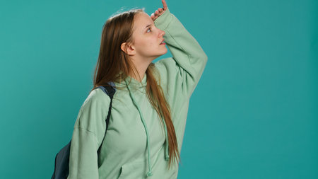Young woman holding backpack looking around for something, doing hand gesture, isolated over studio background. Radiant teenager with good eyesight seeking things, camera Aの写真素材