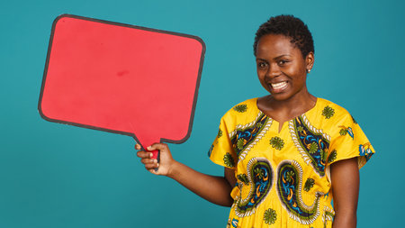 Smiling person showing a red isolated speech bubble in studio, holding a cardboard icon for a commercial advertisement. Young woman in tribal attire presenting an empty carton sign. Camera A.の写真素材