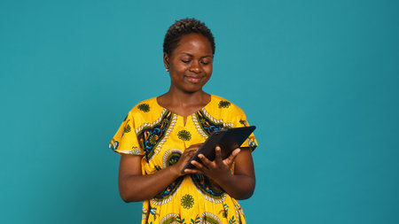 Smiling happy person checking internet websites on her tablet in studio, wearing african ethnic clothes with tribal elements. Confident pleased girl browses online social media sites. Camera A.の写真素材