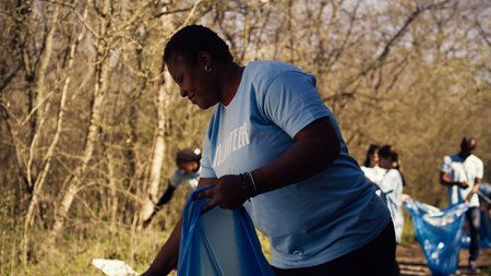 African american activist doing litter cleanup to fight illegal dumping, protecting the natural environment with garbage disposals. Woman working on collecting rubbish from the forest area. Camera B.の写真素材