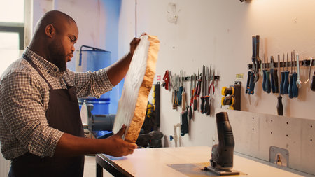 BIPOC man inspecting wood in assembling shop, putting protection equipment on and picking tool from rack. African american cabinetmaker picking lumber block, preparing to do piling on it, camera Bの写真素材