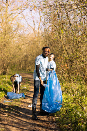 African american activist collecting garbage junk and plastic waste from the woods, storing rubbish in a trash bag and cleaning the natural environment. Environmentalist gathering litter.の写真素材