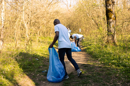 African american man volunteering to clean the forest habitat, collecting junk and plastic waste to help preserve the natural environment. Activist grabbing trash and recycling in disposal bags.の写真素材