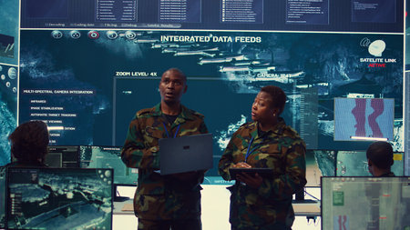 Sergeant major and female soldier checking real time field data in command post monitoring room, examining missions and operations. Team of troops interprets satellite imagery. Camera B.の写真素材
