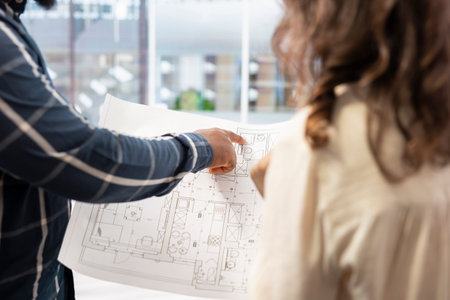 Real estate expert presenting office space layout to woman using blueprints and architectural plans, evaluating the property for potential startup relocation and investment. Modern workspace.の写真素材