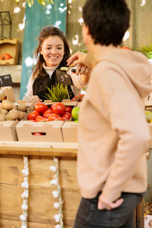 Smiling merchant gives female client fruit sample for tasting and selling healthy at local farmers market. Friendly woman farm stand owner offering consumer to try eco products before purchasing it.の写真素材