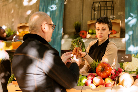 Small business run by woman selling organic bio fruits and vegetables at a local farmers market stand. Food market vendor shows a senior customer fresh sustainable produce at outdoor farming store.の写真素材