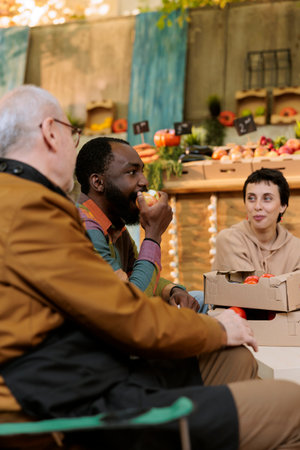 Customers have fun with farmers market stand owner, male client bites an apple at food tasting festival in the autumn. Multiracial people eating organic bio produce and sampling fruits and vegetables.の写真素材