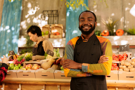 Portrait of young black man wearing an apron, standing near food marketplace with arms crossed. African american vendor at farmers market booth waiting for customers, looking at camera.の写真素材