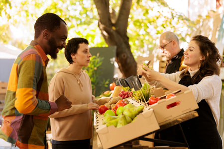 Multicultural couple man and woman purchasing seasonal natural produce at farmers market, looking at bio products. Smiling vendor standing behind fruits and vegetables stand, selling healthy food.の写真素材