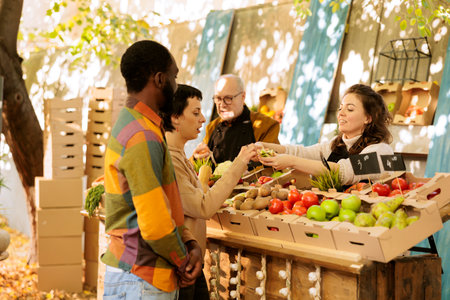 Female merchant offering samples to customers while selling fresh bio fruits and vegetables at farmers market. Young multiracial family trying seasonal organic produce, visiting food fair festivalの写真素材