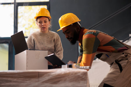 Diverse warehouse coworkers preparing cu clients orders using cardboard boxes, checking delivery details on laptop in storage room. African american worker with protective overall working at inventoryの写真素材
