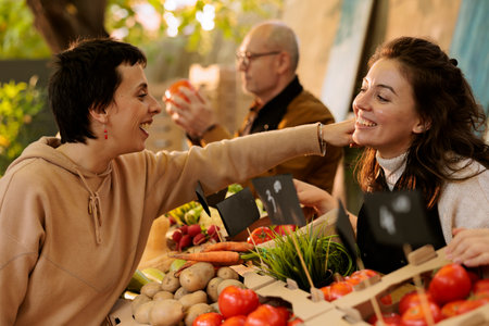 Cheerful local vendor greets frequent shopper at food market stand while showing fresh organic produce. Pleasant farmer hugging customer and selling locally grown produce at harvest fair festival.の写真素材
