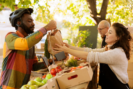 Female merchant giving packaged order of fresh produce to african american male courier, helping with fruits and vegetables delivery. Friendly woman farmer preparing organic eco products.の写真素材