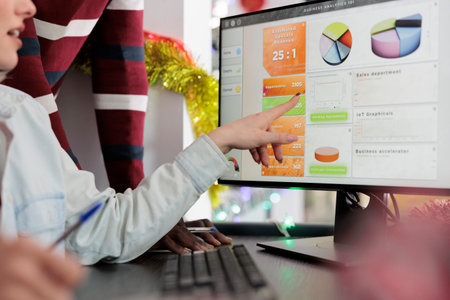 Businesswoman in Christmas decorated office showing coworkers key data point on computer screen needed for marketing insight. Employees working together on finishing project before impending deadlineの写真素材