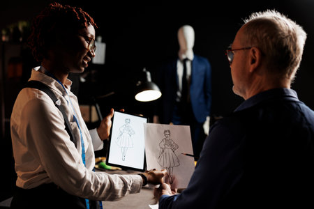 African american tailor apprentice and elderly master dressmaker comparing sketch drawing on tablet and paper, trying to design the perfect dress. Couturiers working on client comissionの写真素材