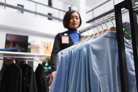 Blue casual shirts hanging on apparel rack while asian woman seller standing in mall clothing store selective focus. Stylish garment on hangers in department center boutique closeupの写真素材