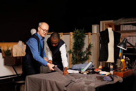 Experienced dressmakers cutting fabric, manufacturing stylish sleek business clothing in refined masterful craftmanship. Tailors working with textile materials in atelier shop, creating bespoke attireの写真素材