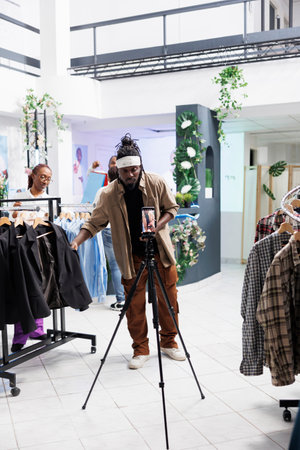 African american social media influencer promoting formal wear collection on smartphone camera in clothing store. Blogger showing jackets while filming live video for followers in boutiqueの写真素材