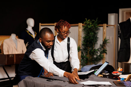 Refined masterful tailor team cutting fabric for bespoke modern fashion collection in dark studio. Elegant dressmakers manufacturing attire following dresses sketchesの写真素材