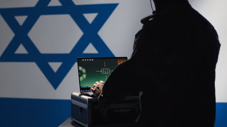 Israel Defense Forces dispatcher looking at surveillance transmission on laptop, relaying data to troops using headphones microphone, isolated over israeli flag background, camera Aの写真素材