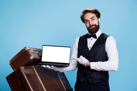 Bellboy showcases white screen on pc presenting hospitality industry professionalism, studio background. Doorman advertising multitasking ability to provide assistance and show blank display.の写真素材