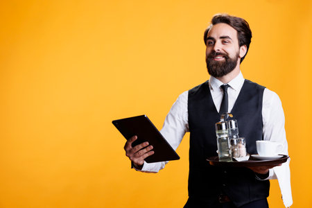Young butler handing out bill on tablet, providing excellent five star service and assistance on camera. Professional employee using device while he holds tray platter in hand.の写真素材