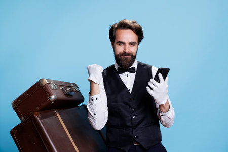 Smiling bellhop on mobile phone, feeling happy about good news on internet sites in studio. Positive remarkable doorkeeper with gloves acting excited and positive on camera.の写真素材