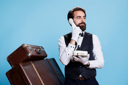 Classy bellhop interacts with tourists about facilities as they make calls on landline phone. Professional competent doorman using wired telephone to take inquiries for aristocratic job.の写真素材