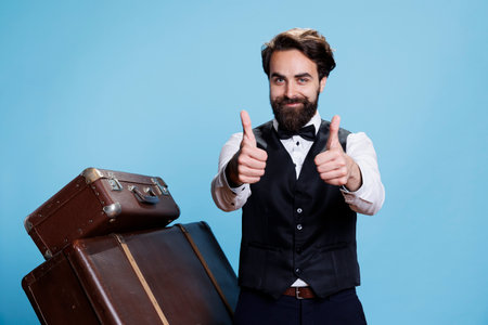 Hotel employee does like sign on camera, acting content and cheerful against blue studio background. Stylish elegant bellboy shows thumbs up in agreement, positive approval gesture.の写真素材